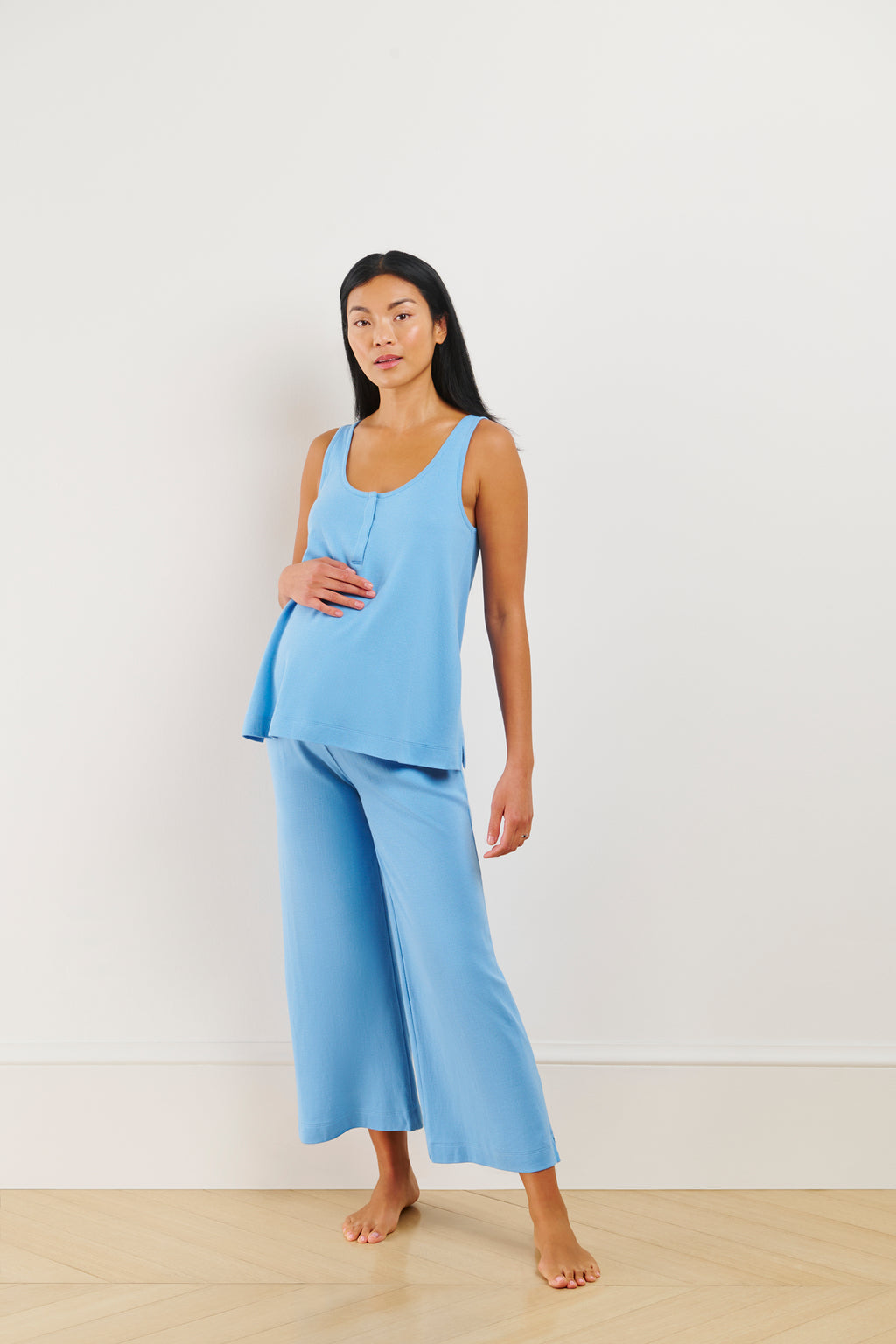 A pregnant woman stands against a plain white wall, wearing the Pointelle Maternity Wide Leg Tank Set in Serene Blue. She gently holds her belly with one hand and looks calmly at the camera.