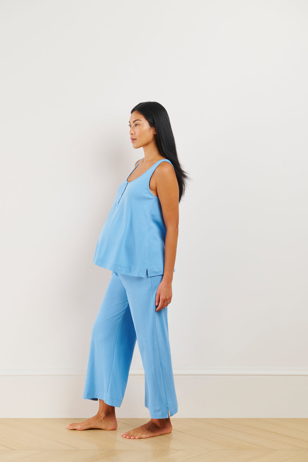 A pregnant woman wears the Pointelle Maternity Wide Leg Tank Set in Serene Blue, featuring a loose-fitting tank top and matching wide-leg pants, while standing barefoot against a plain white wall.