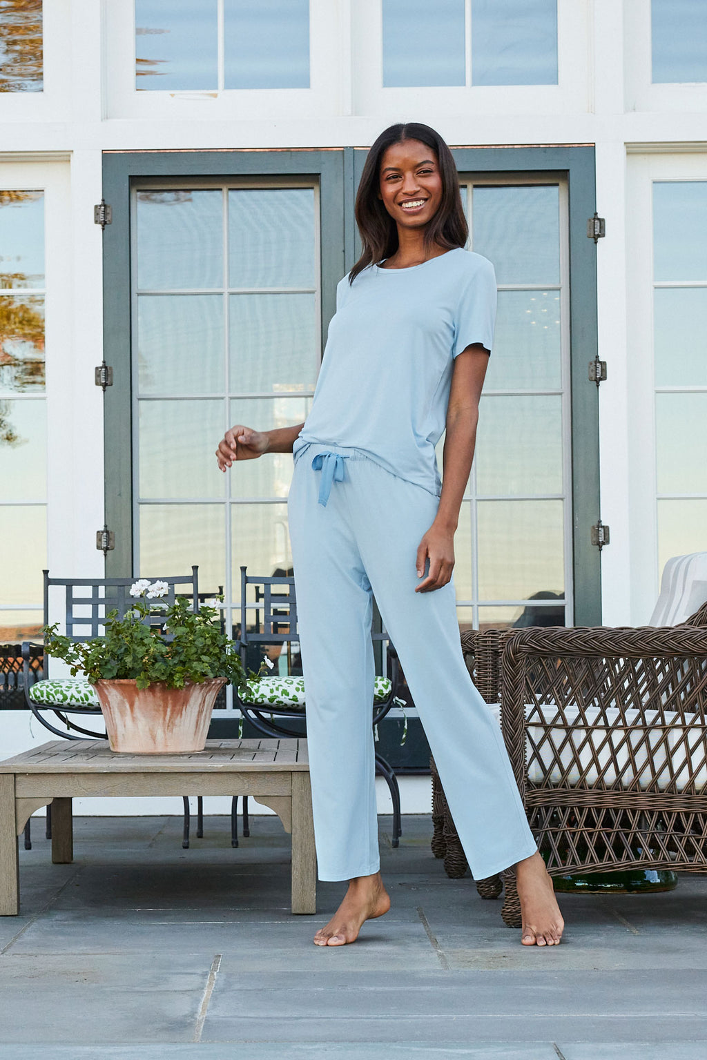 Wearing the DreamModal Ribbon Short-Long Set in French Blue, a woman stands on a patio with potted plants, outdoor chairs, and double doors behind her, smiling at the camera.
