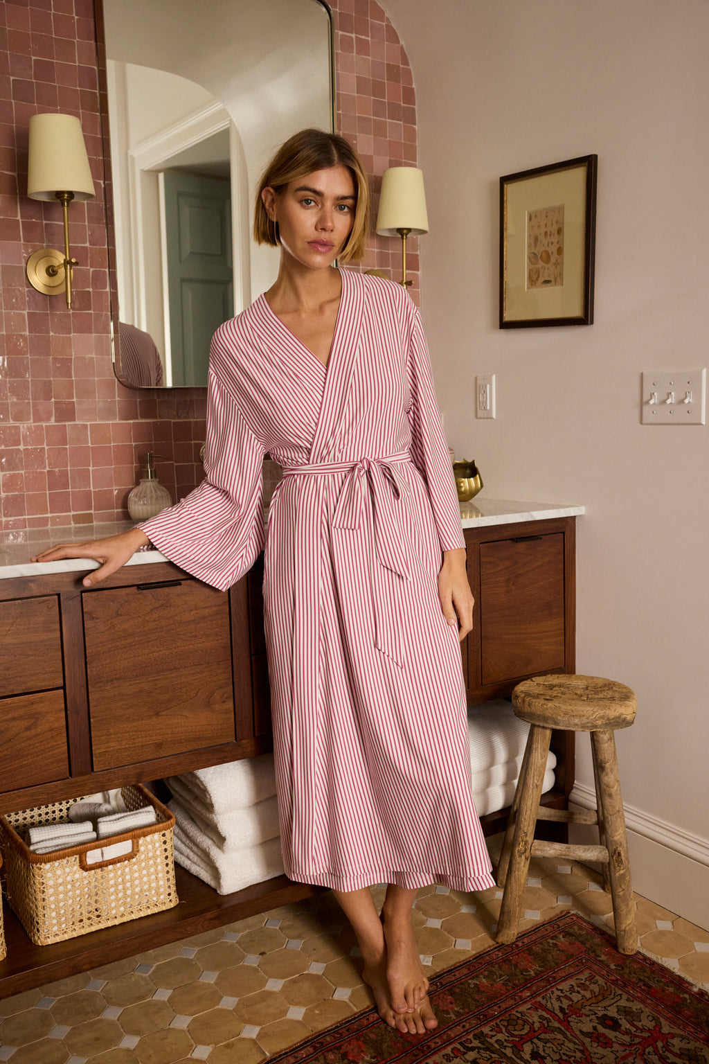 A woman wearing the DreamModal Kimono Robe in Camellia Pencil Stripe stands in a bathroom with pink tiled walls, wooden cabinets, a round mirror, and a small wooden stool.