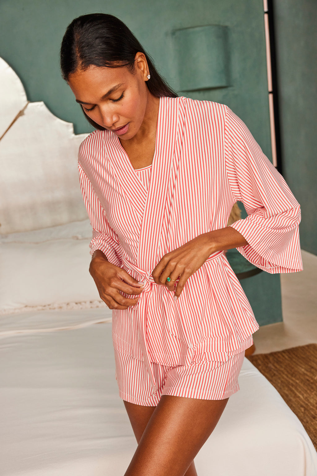 A woman stands by a bed, tying the waist belt of her DreamModal Kimono Shorts Bundle in Terrace Pencil Stripe. The room features modern, serene decor with a green wall in the background.