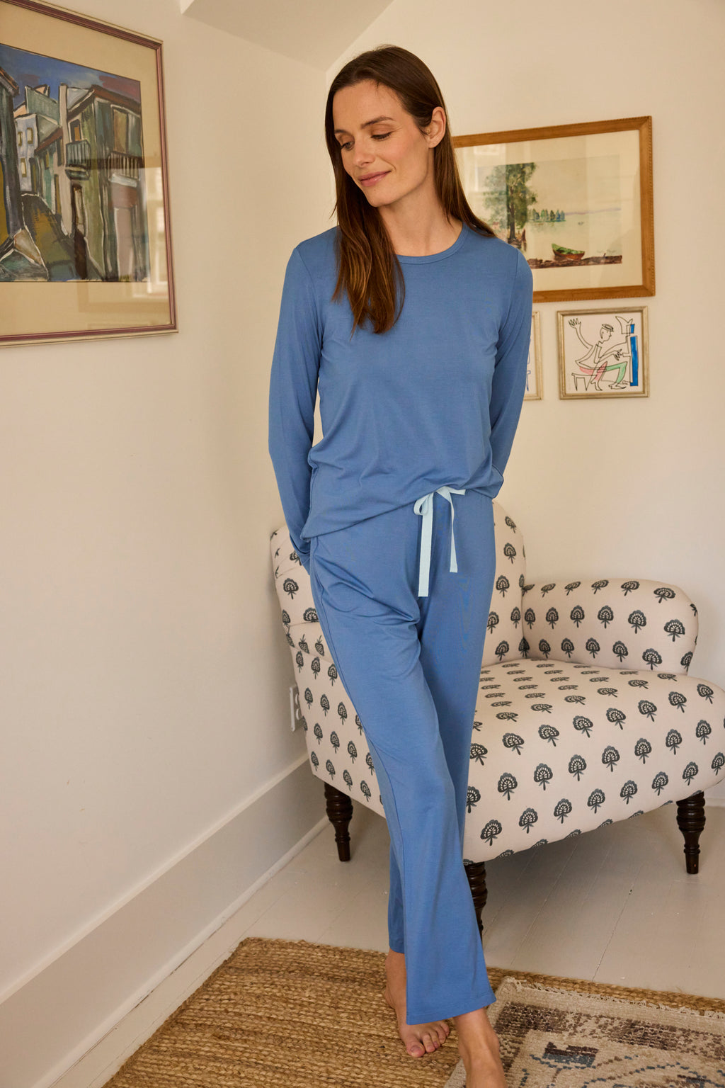 A woman wearing the DreamModal Ribbon Long-Long Set in Seaport stands smiling on a woven rug in a cozy room, with a patterned armchair and framed artwork on cream-colored walls behind her.