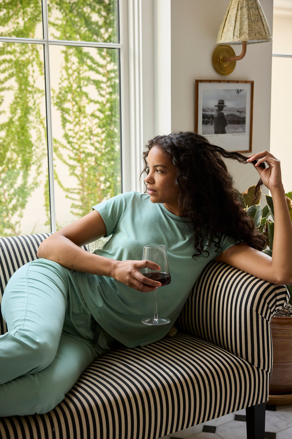 A woman in a Granite Green DreamModal Ribbon Short-Long Set relaxes on a striped sofa, holding red wine and gazing out at greenery. A framed photo and plants create a charming backdrop.
