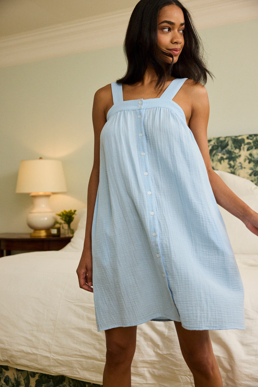 A woman stands indoors wearing a sleeveless Hamptons Nightgown in French Blue, made from soft cotton modal. She’s in a bedroom with a bed, white bedding, and a bedside lamp—ideal comfort for hot sleepers.