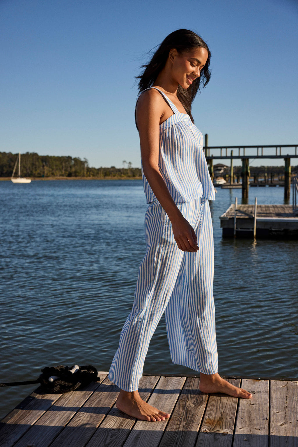 A woman in the Hamptons Pants Set in Sail Blue Awning Stripe stands on a wooden dock by the water, with a sailboat and trees behind her on a sunny day—the airy gauze fabric is perfect for hot sleepers.