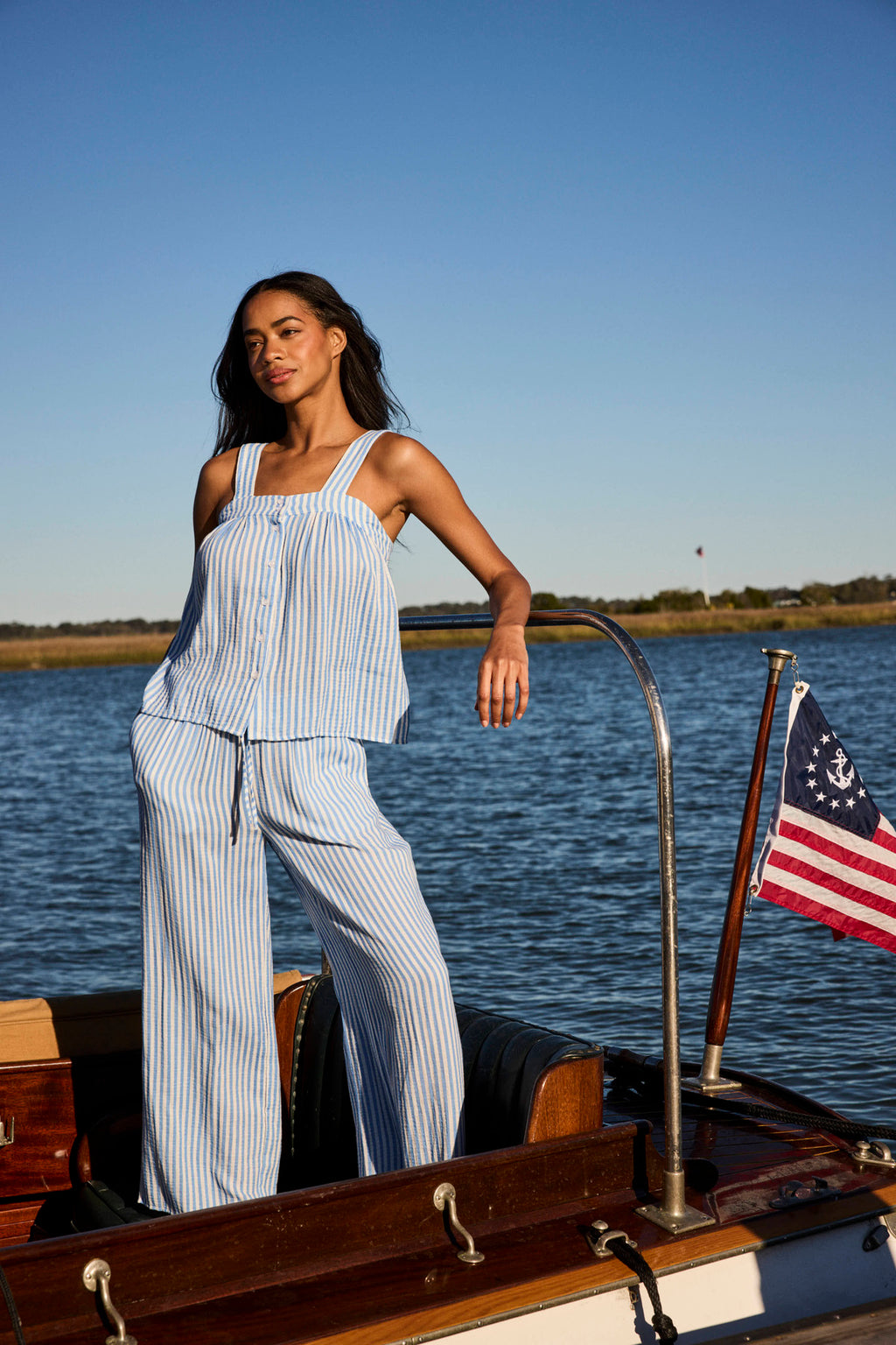 A woman in the Hamptons Pants Set in Sail Blue Awning Stripe stands on a wooden boat with water, greenery, and an American flag at the stern. The breezy gauze fabric keeps her cool on warm days.
