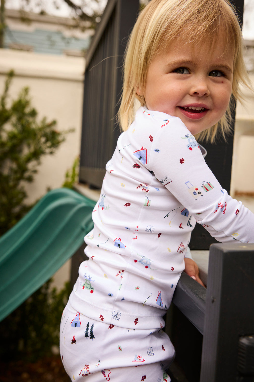 A child in the Kids Long-Long Set in Alpine Adventure—soft Pima cotton pajamas with colorful prints—smiles while climbing a playground structure beside a green slide, with shrubs and a fence in the background.