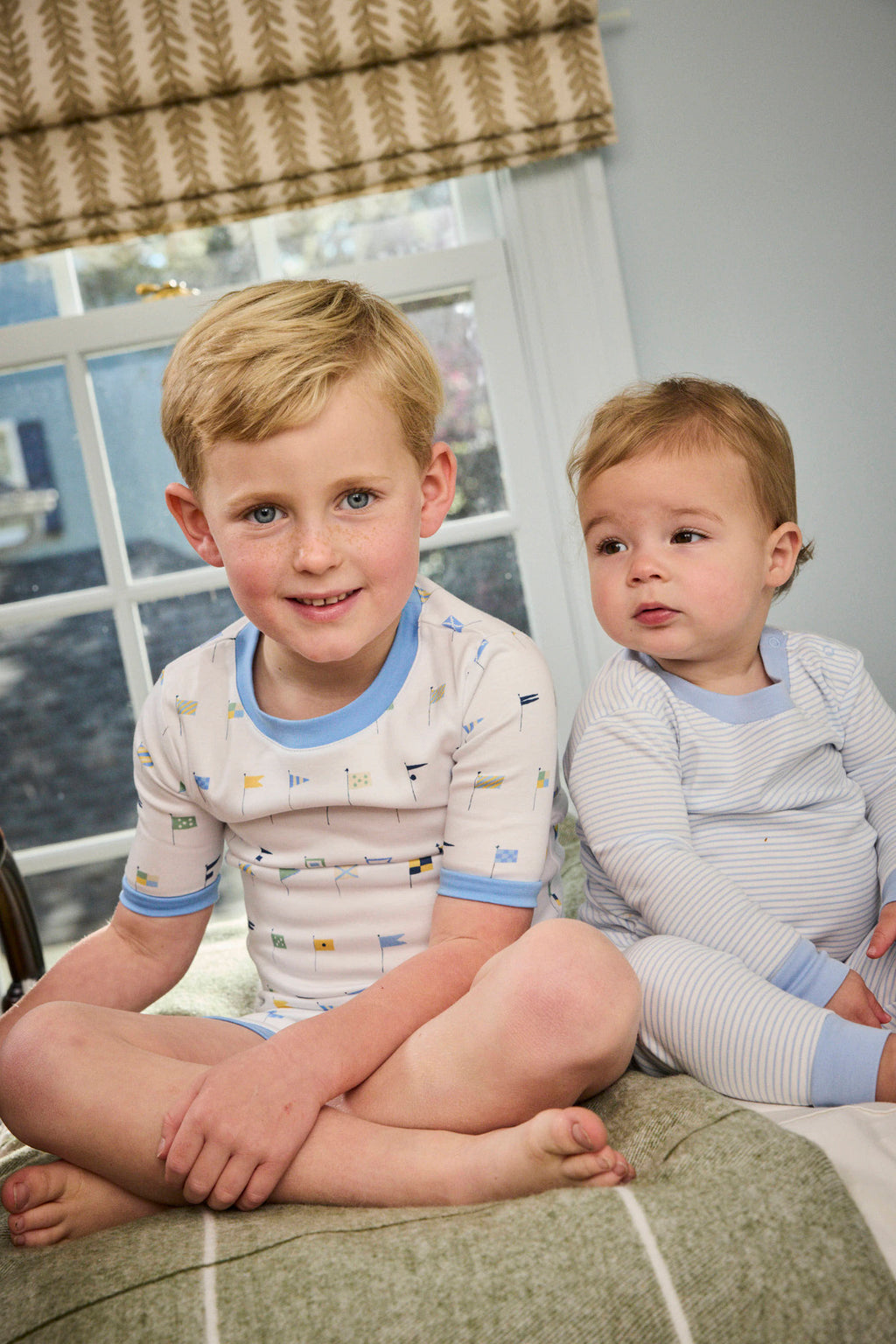 Two young children sit on a bed near a window. The older child wears the Kids Shorts Set in Baltic Blue Nautical Flags and smiles at the camera, while the younger one, dressed in striped pajamas, looks off to the side.