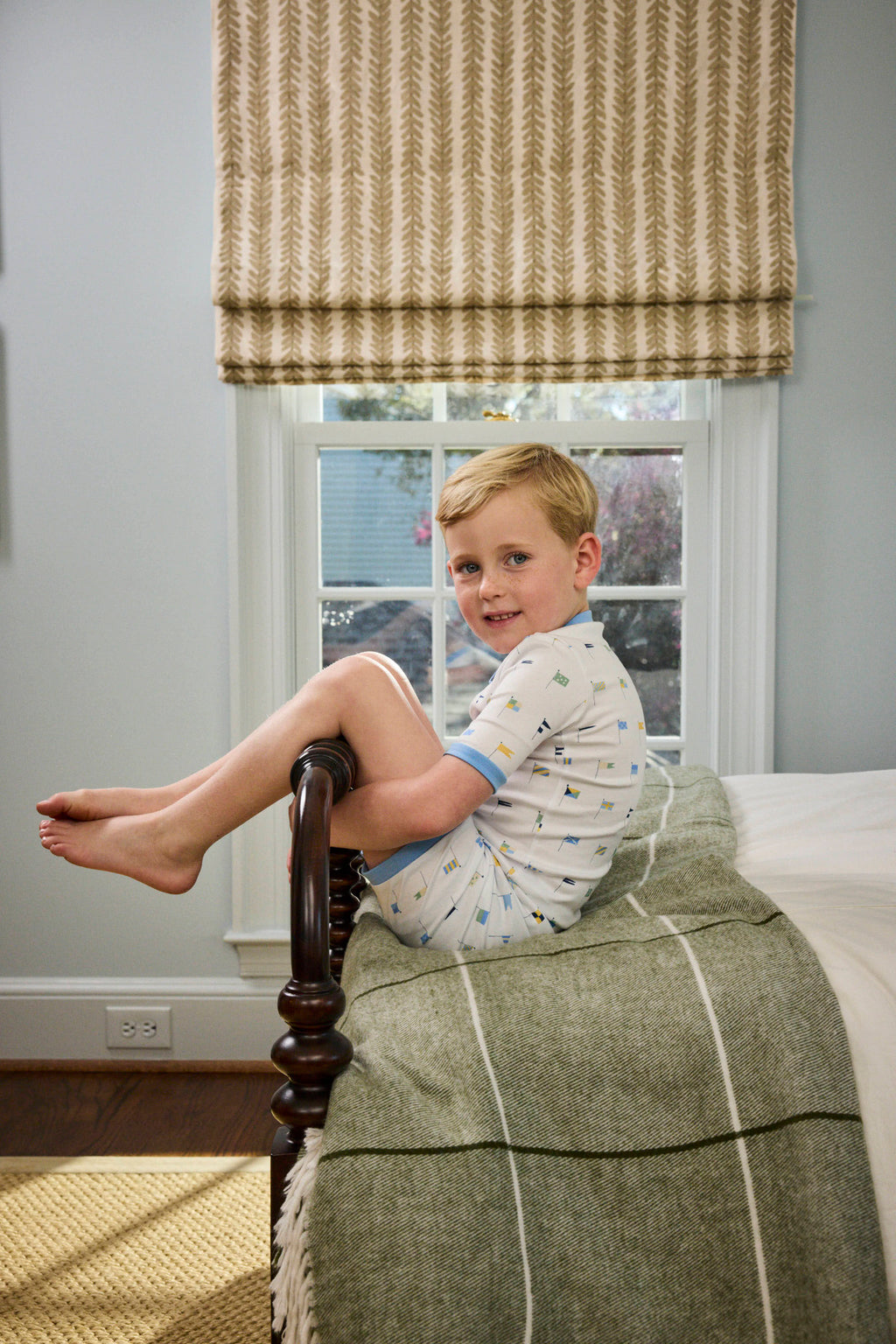 A young boy in the Kids Shorts Set in Baltic Blue Nautical Flags sits sideways at the end of a wooden bed, smiling at the camera. Behind him are a window with a beige patterned shade and a green blanket on the bed.