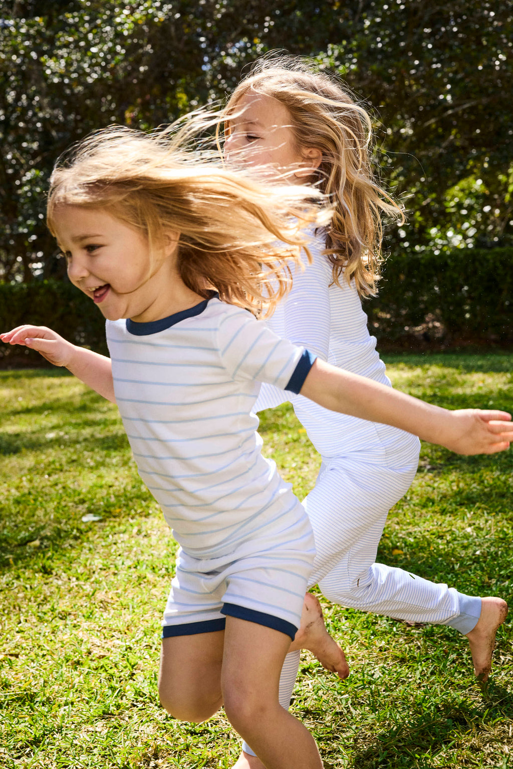 Two young children run joyfully on green grass in the sunshine, both wearing the Kids Shorts Set in Marine. Navy trim accents their light-colored Pima cotton outfits, with trees and shrubs visible in the background.