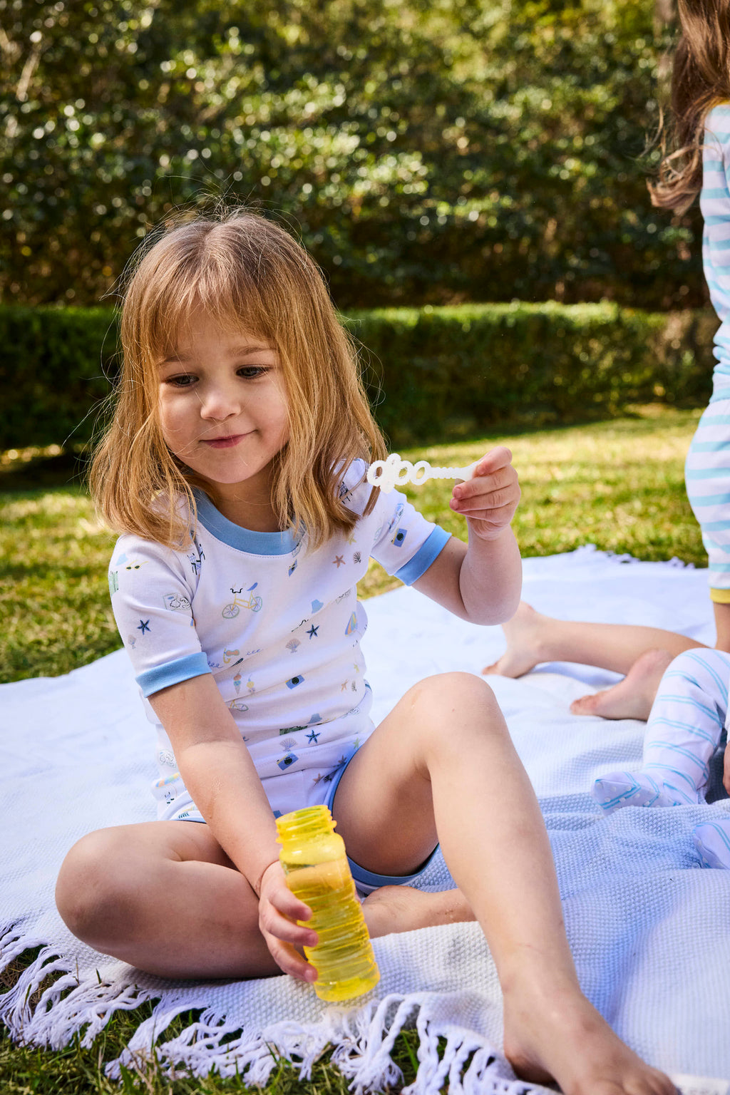 A young girl sits on a white blanket outdoors, holding a bubble wand and bottle of solution. She wears the Kids Shorts Set in Surfside with small blue designs and looks content while playing on the grass.