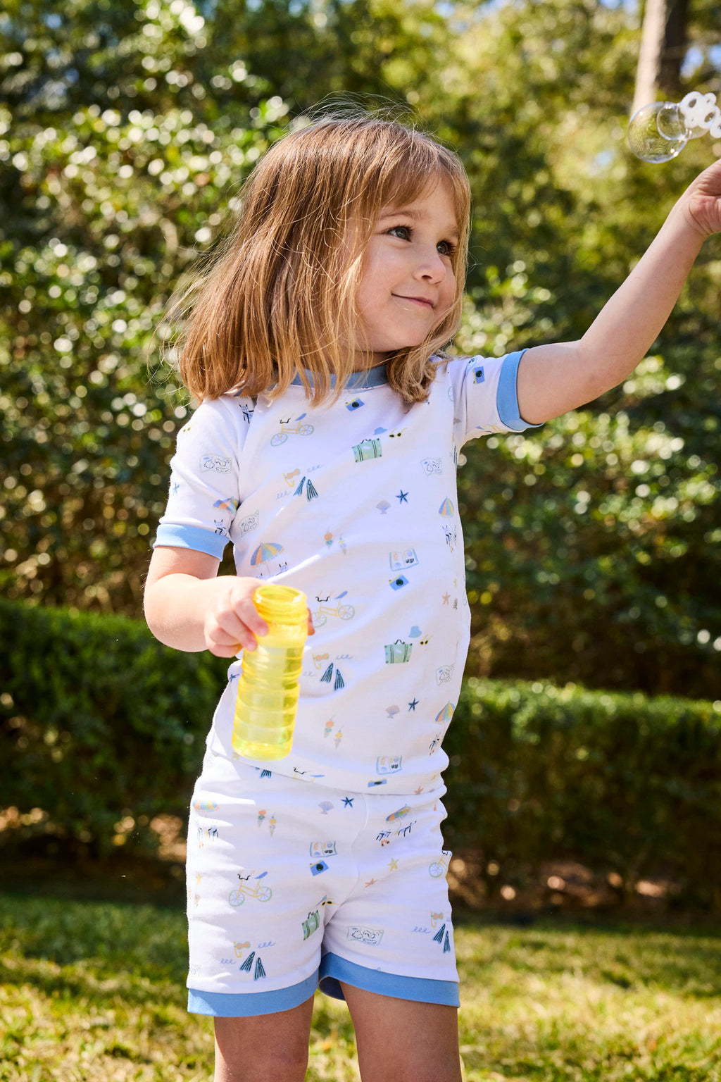 A young child wearing the Kids Shorts Set in Surfside with a beach print stands outside, smiling and blowing bubbles. She holds a yellow bubble bottle, surrounded by lush green bushes and trees in the background.