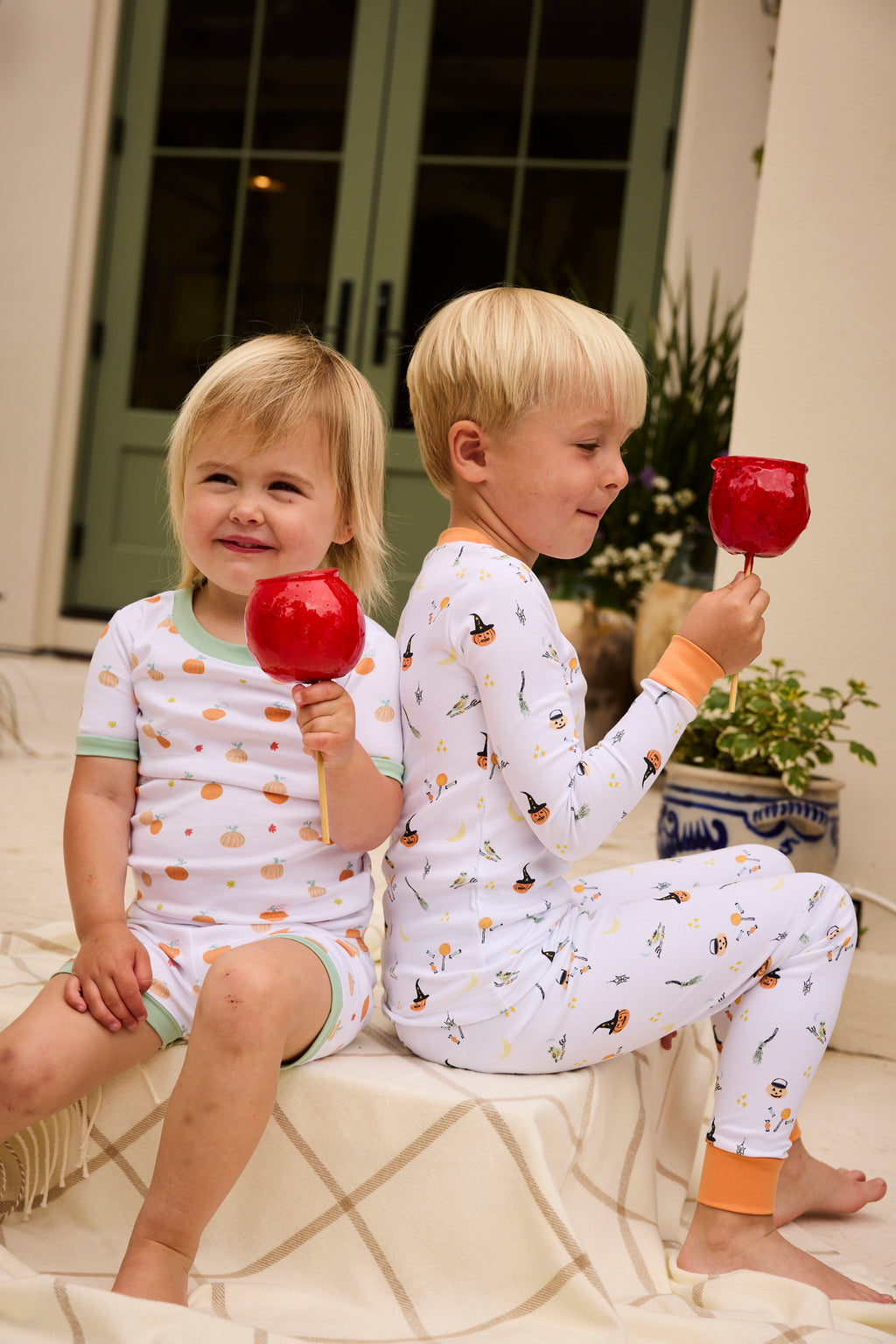 Two kids sit back-to-back on a blanket outdoors, each holding a red candied apple. They wear the Kids Long-Long Set in Trick or Treat, featuring white Halloween pajamas with colorful patterns. Green double doors and potted plants are behind them.
