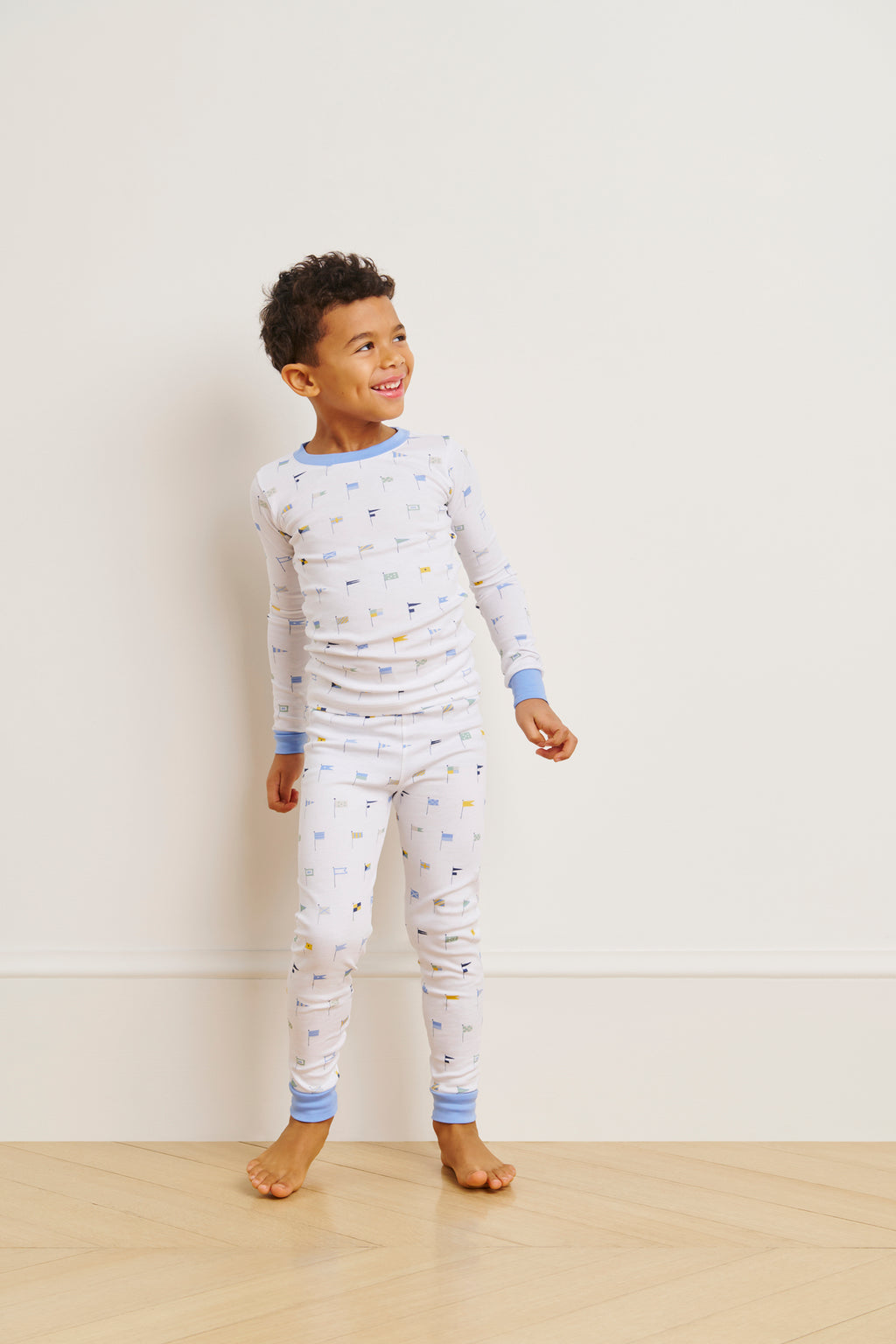 A barefoot young child smiles to the side, wearing the Kids Long-Long Set in Baltic Blue Nautical Flags, crafted from soft Pima cotton with blue cuffs, on a wooden floor against a plain light wall.