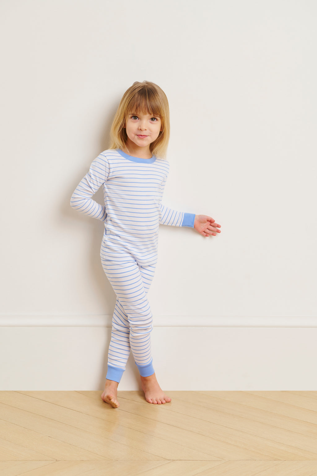 A young child stands on a wooden floor against a white wall, wearing the Kids Long-Long Set in Baltic Blue, made from soft Pima cotton.