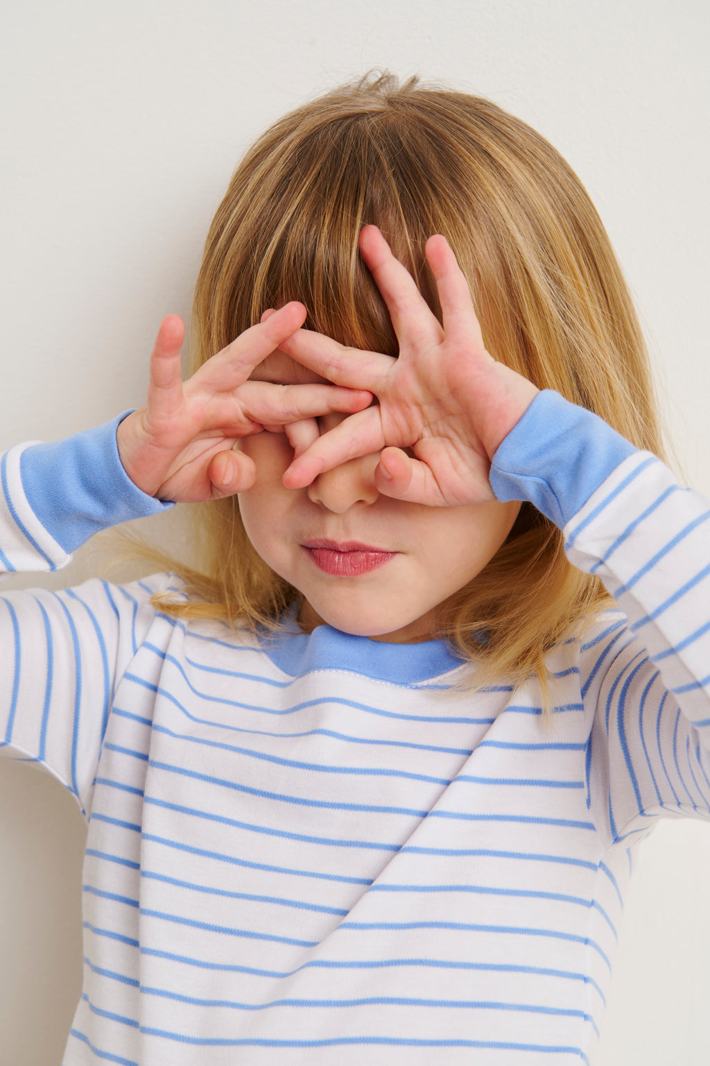 A young child in the Kids Long-Long Set in Baltic Blue playfully covers their eyes and peeks through their fingers, standing against a plain white background.