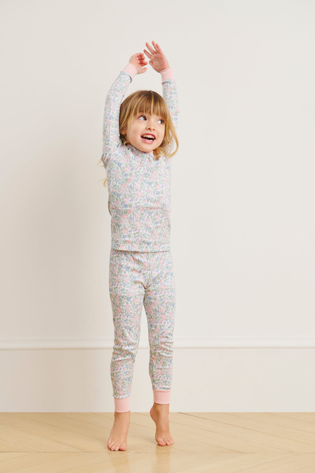 A young child in the Kids Long-Long Set in English Rose Elizabeth Floral, made from 100% Pima cotton, stands on a wooden floor smiling and stretching with arms raised overhead against a plain light-colored wall.