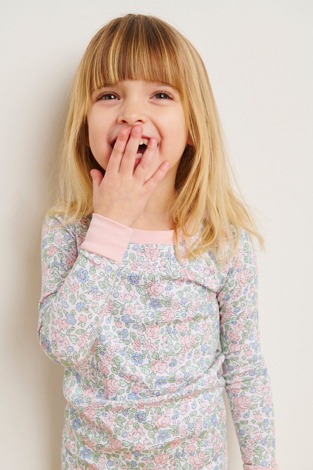 A young girl with bangs smiles with her hand over her mouth, wearing the Kids Long-Long Set in English Rose Elizabeth Floral, made from 100% Pima cotton, standing against a light-colored wall.
