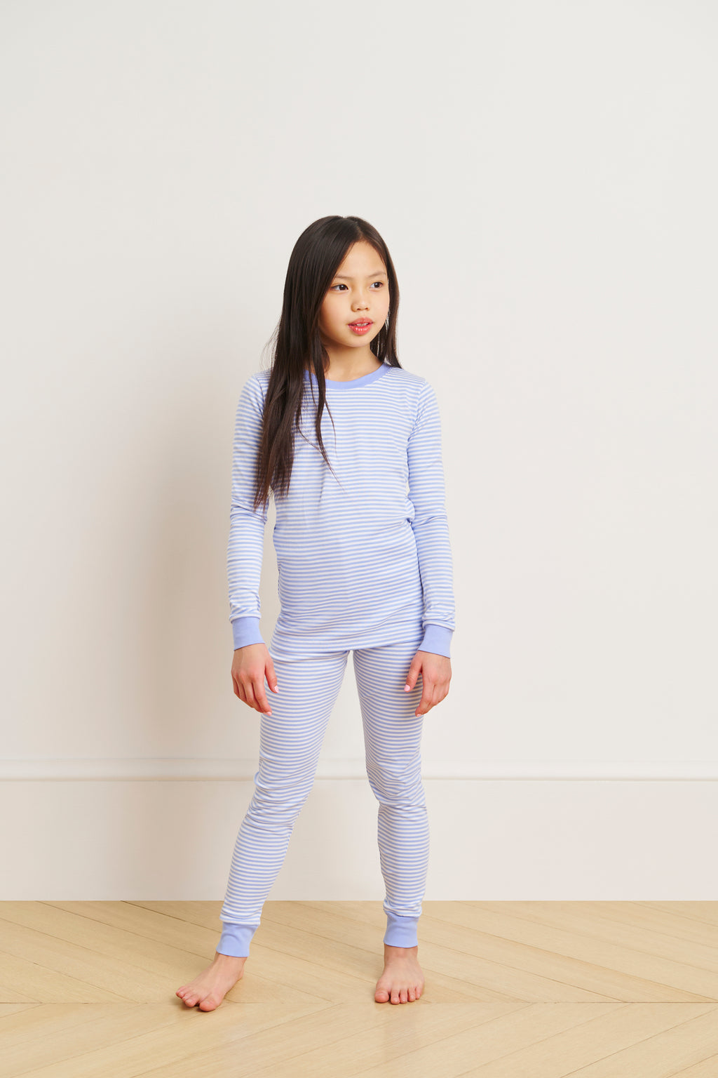 A young girl stands on a wooden floor in the Kids Long-Long Set in Hydrangea, featuring soft Pima cotton pajamas with long sleeves and pants, against a plain white wall.