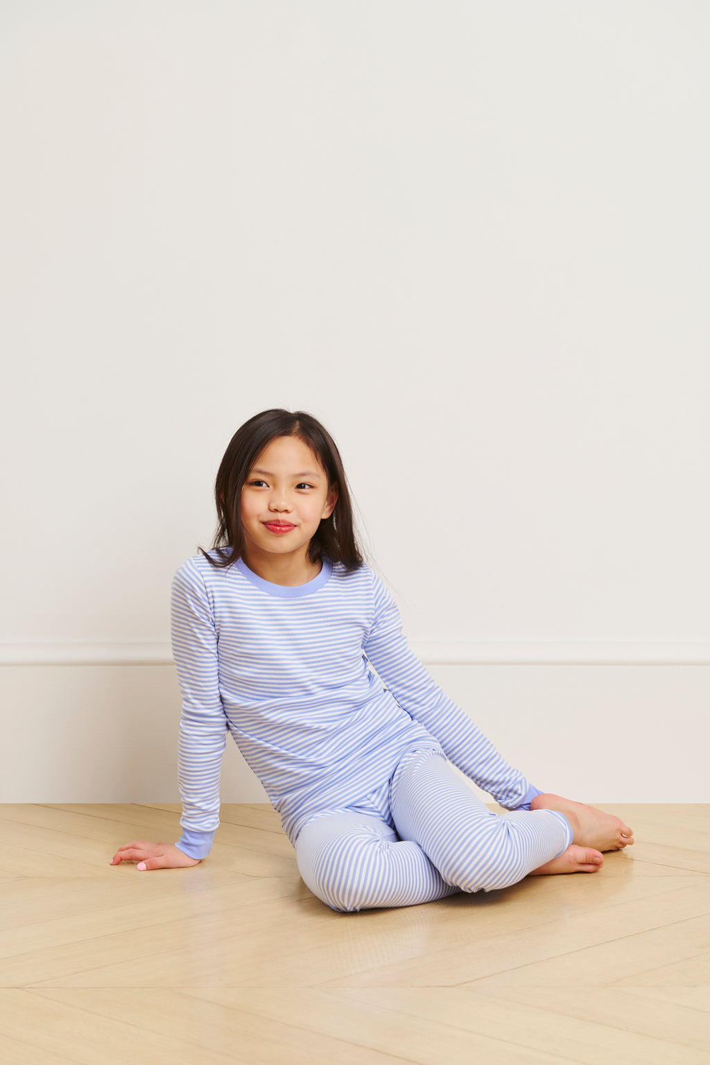 A young child sits relaxed on a wooden floor against a light wall, wearing the Kids Long-Long Set in Hydrangea, made from soft Pima cotton. One hand rests on the floor and their legs are stretched out.