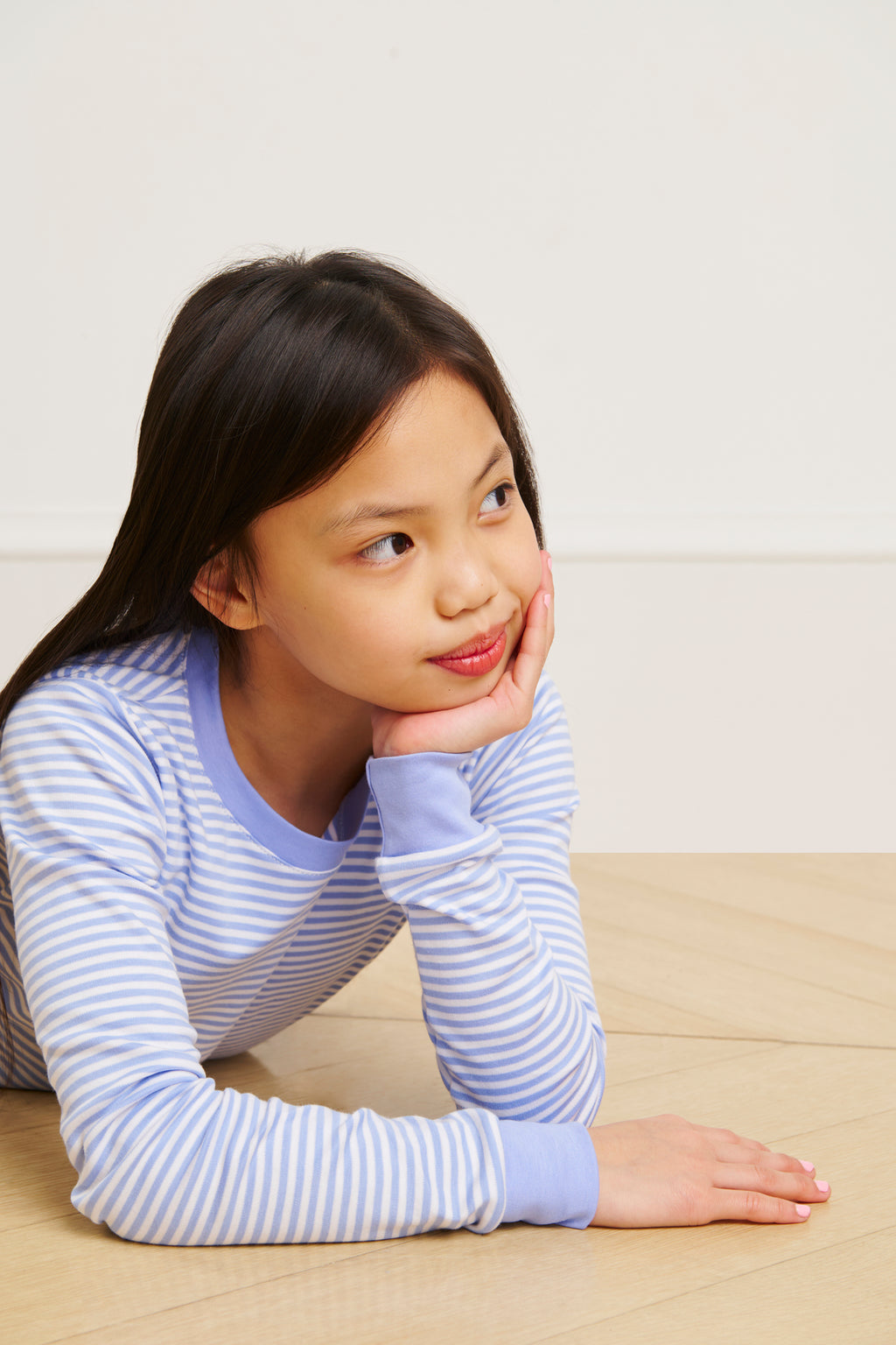 A young girl wearing the Kids Long-Long Set in Hydrangea, made of soft Pima cotton, lies on a wooden floor and gazes thoughtfully to the side with her chin resting on her hand.