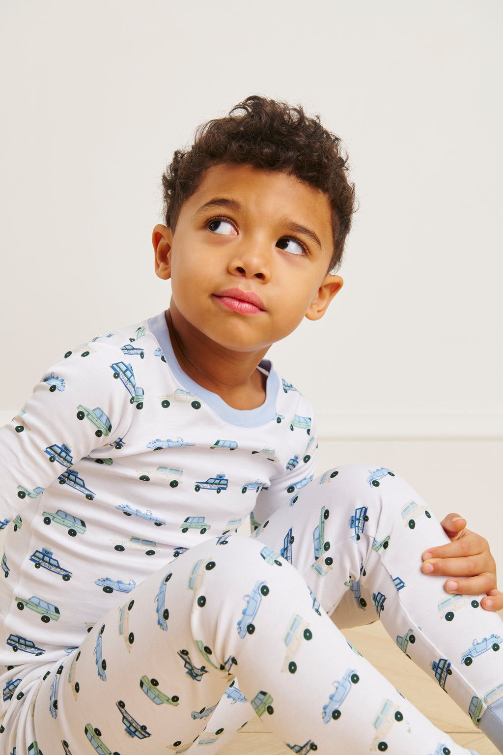 A young child in white Kids Long-Long Set in Off Road Cruisers, featuring blue and green vehicle prints, sits thoughtfully on the floor. The set is crafted from soft Pima cotton and shown against a plain background.