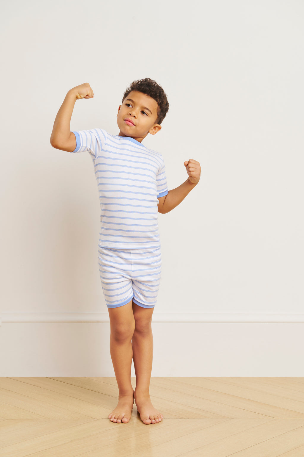 A young boy confidently flexes his arms while wearing the Kids Shorts Set in Baltic Blue Triple Stripe, standing on a wooden floor against a plain, light-colored wall that highlights the comfy Pima Cotton fabric.