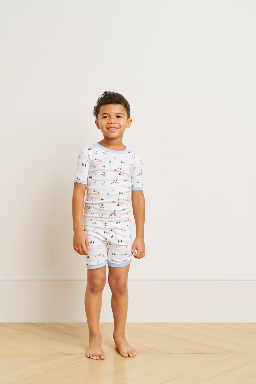 A young boy smiles barefoot on a wooden floor, wearing the Kids Shorts Set in Camp LAKE with colorful animal prints, against a plain light wall. Perfect for summer camp adventures and made from soft Pima cotton.