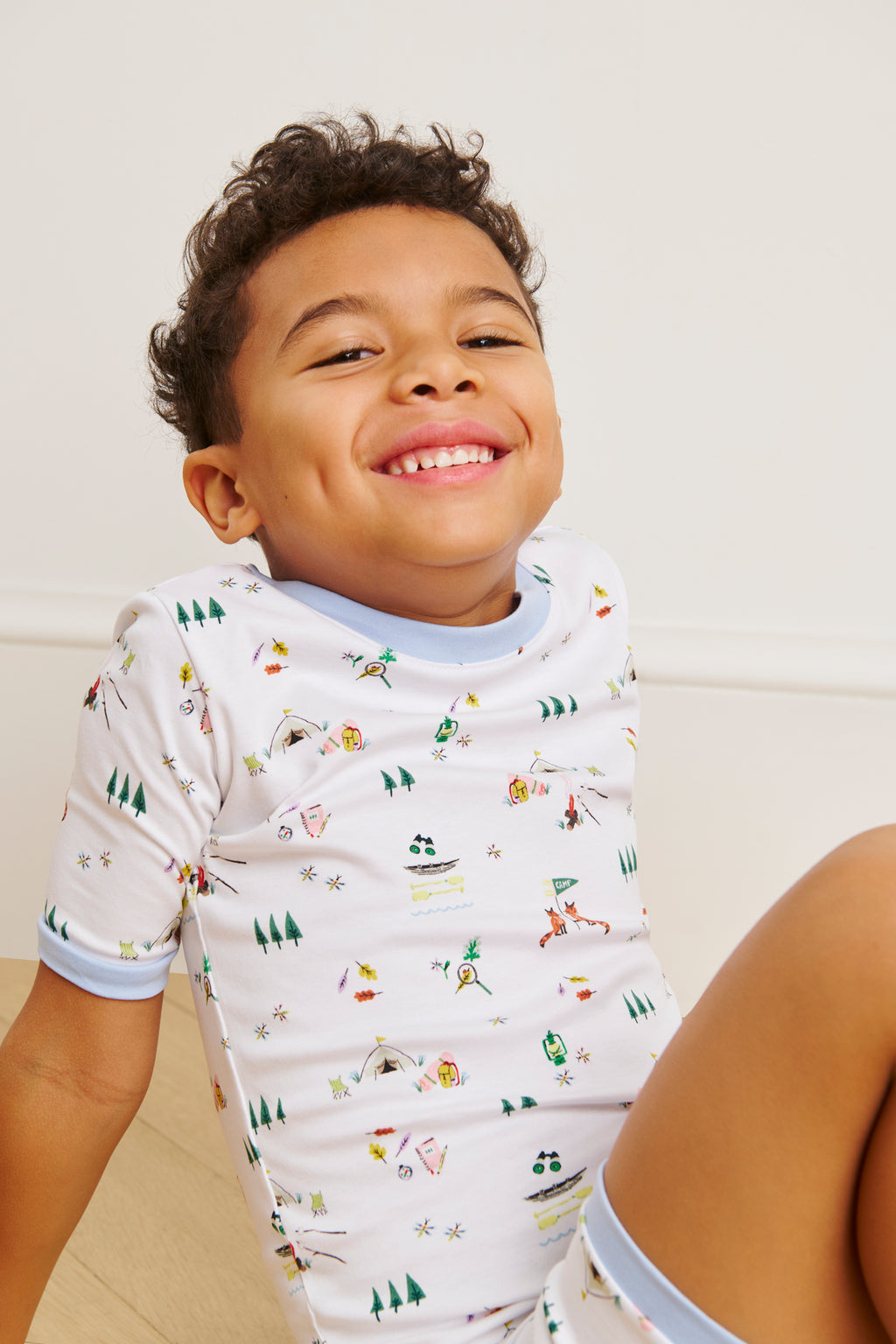 A smiling young child sits on the floor, wearing the Kids Shorts Set in Camp LAKE, featuring soft Pima cotton and playful prints. The outfit is accented with light blue trim, set against a plain, light-colored background.