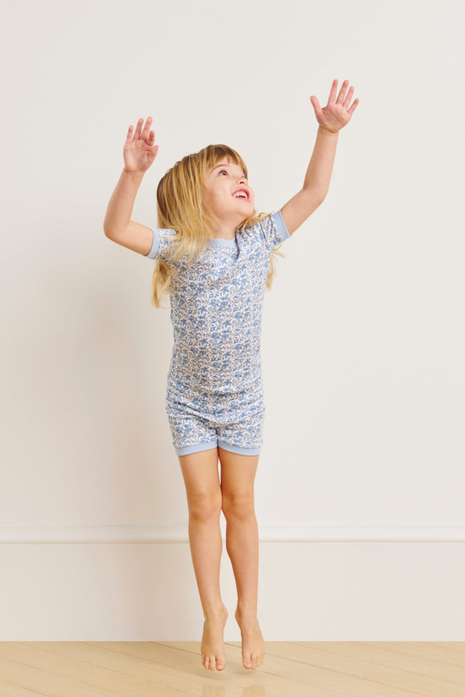 A young child smiles with arms raised, barefoot in the Kids Shorts Set in Delft Blue Garden Floral—made from 100% Pima cotton for ultimate comfort—standing against a light-colored wall.