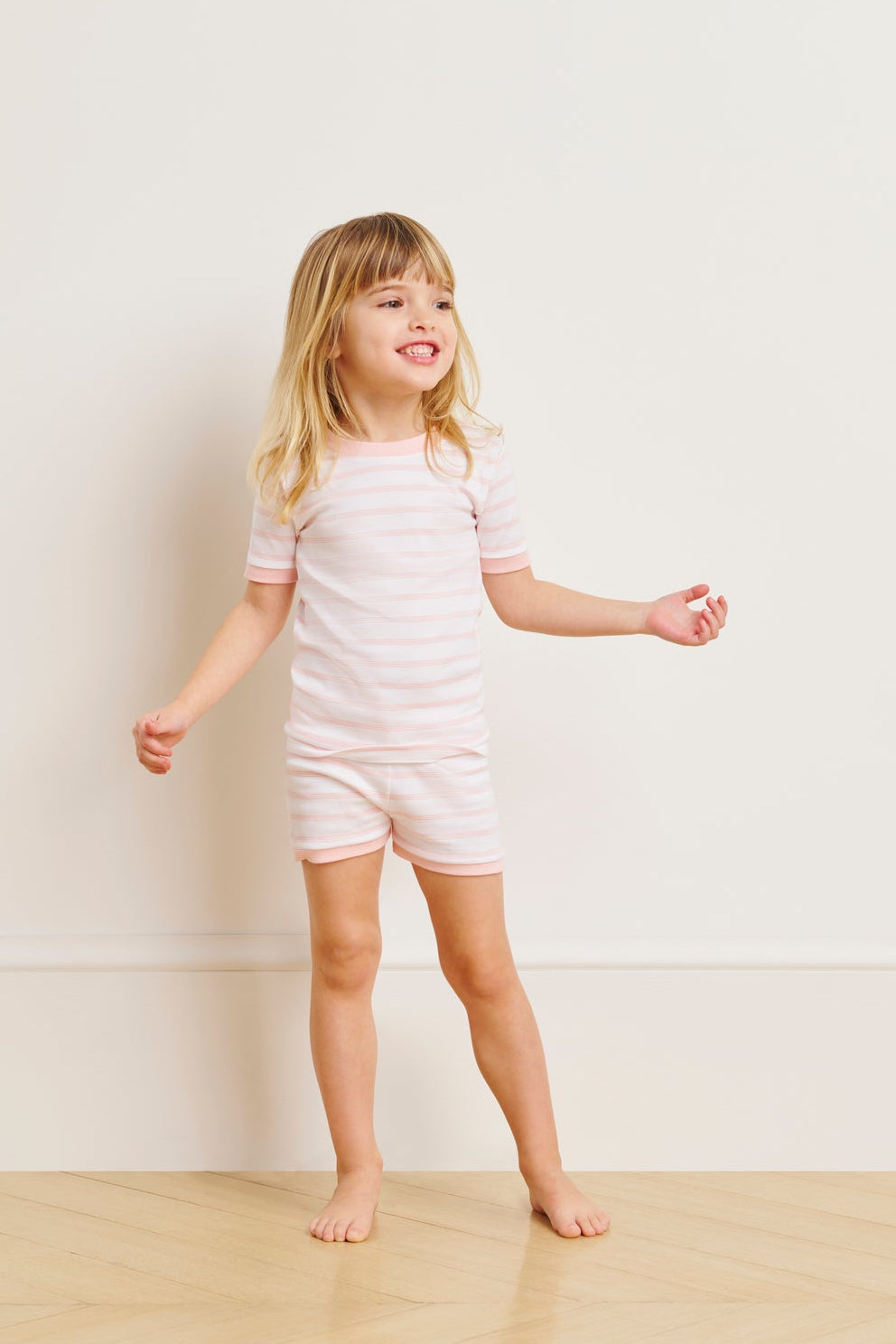A young child smiles barefoot on a wooden floor, wearing the Kids Shorts Set in English Rose Triple Stripe. The background is a plain white wall.