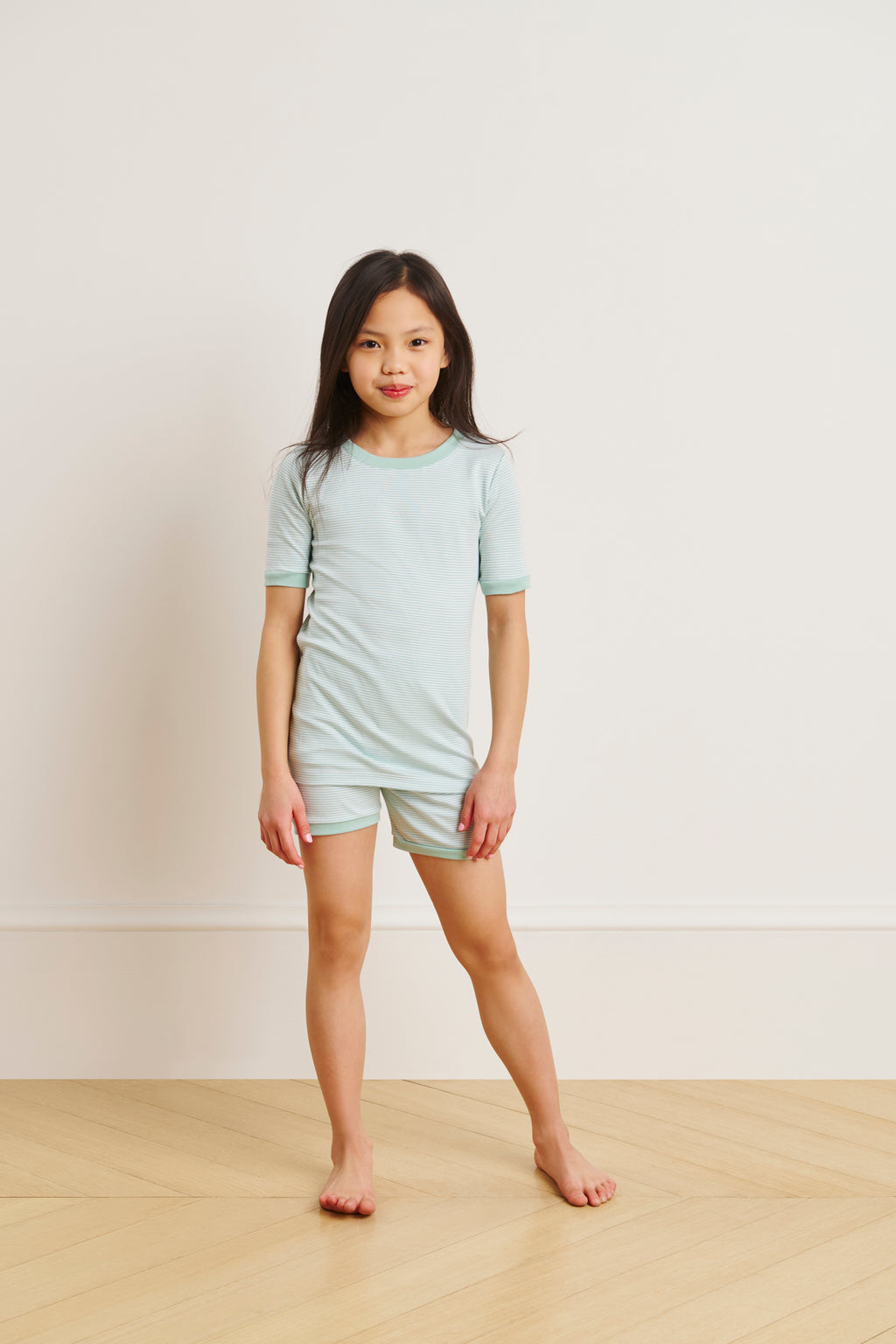 A young girl stands barefoot on a light wooden floor, wearing the Kids Shorts Set in Parisian Green made from soft Pima cotton, with a plain off-white wall in the background.