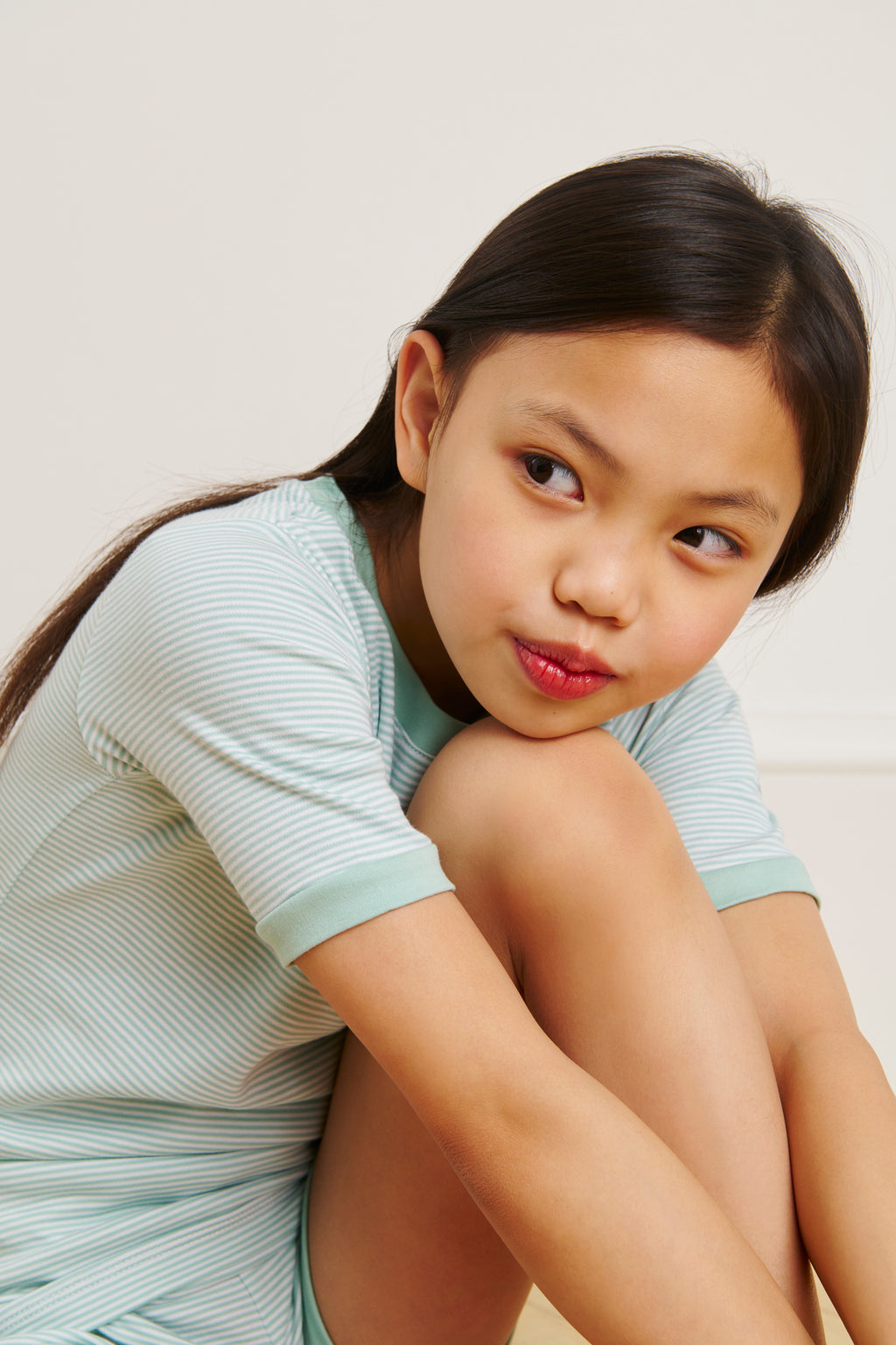A young girl wearing the Kids Shorts Set in Parisian Green sits with one knee up, gazing thoughtfully to her side against a plain background.
