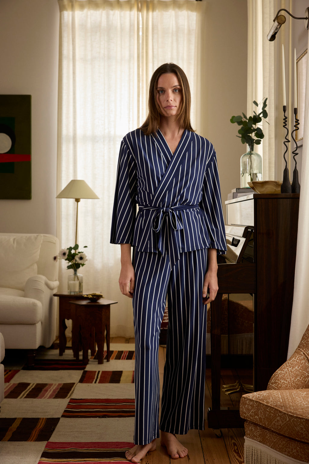A woman in a living room wears the DreamModal Kimono Pajama Set in English Navy Vertical Stripe. She faces the camera with a neutral expression; light curtains, a white sofa, and decorative plants are visible in the background.