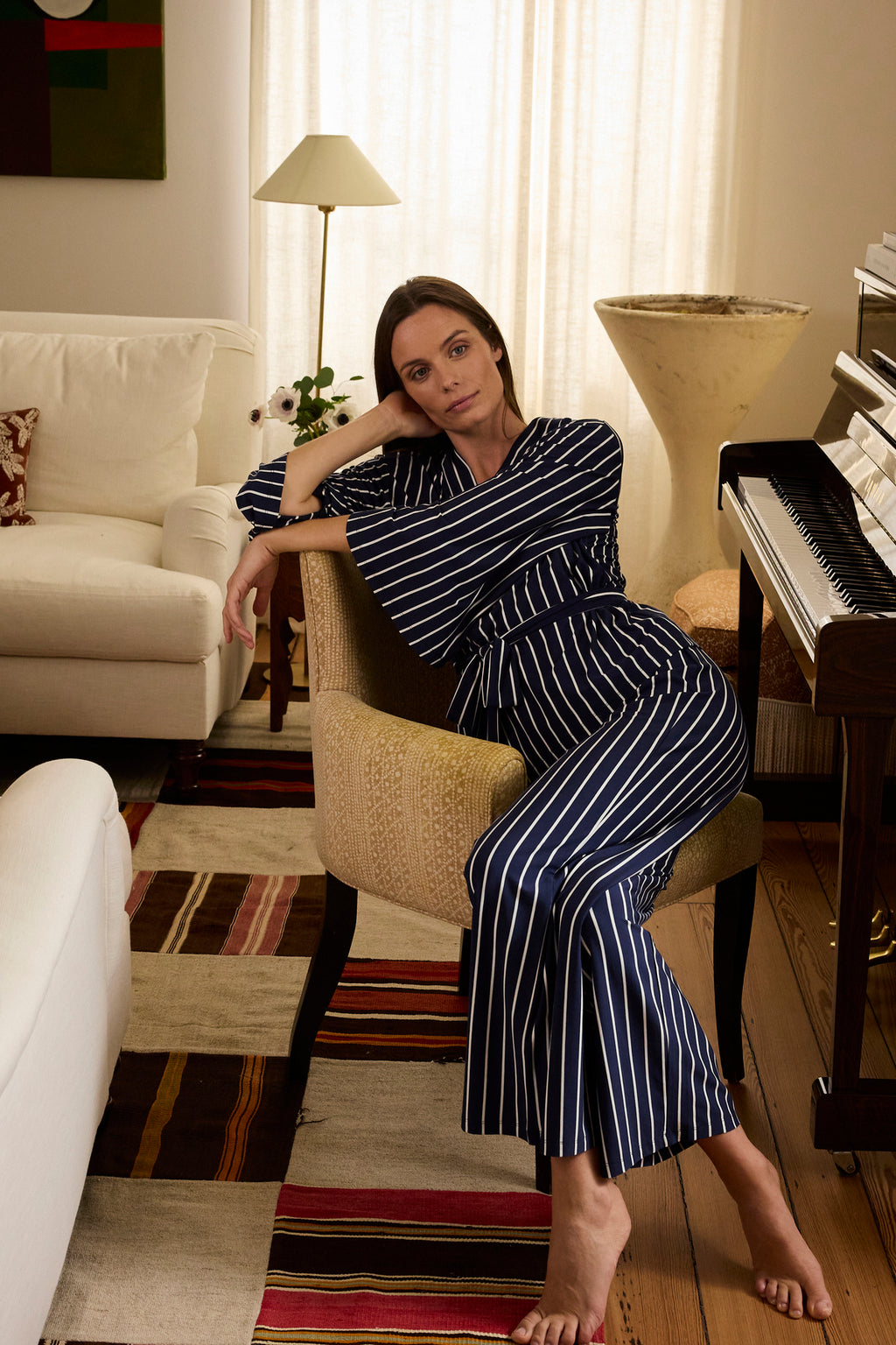 A woman in the DreamModal Kimono Pajama Set in English Navy Vertical Stripe sits on a beige chair, resting her head on her hand in a cozy, softly lit living room with a piano, colorful rug, and cream-colored furniture.