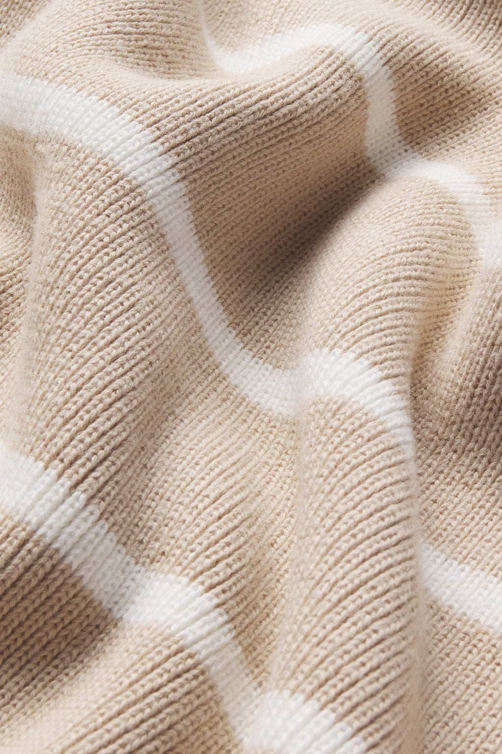 A close-up of the Madeline Cotton Sweater in Driftwood Ivory Stripe, showing beige ribbed knit with intersecting white lines. The soft 100% cotton fabric features a subtly wrinkled texture for a cozy look.