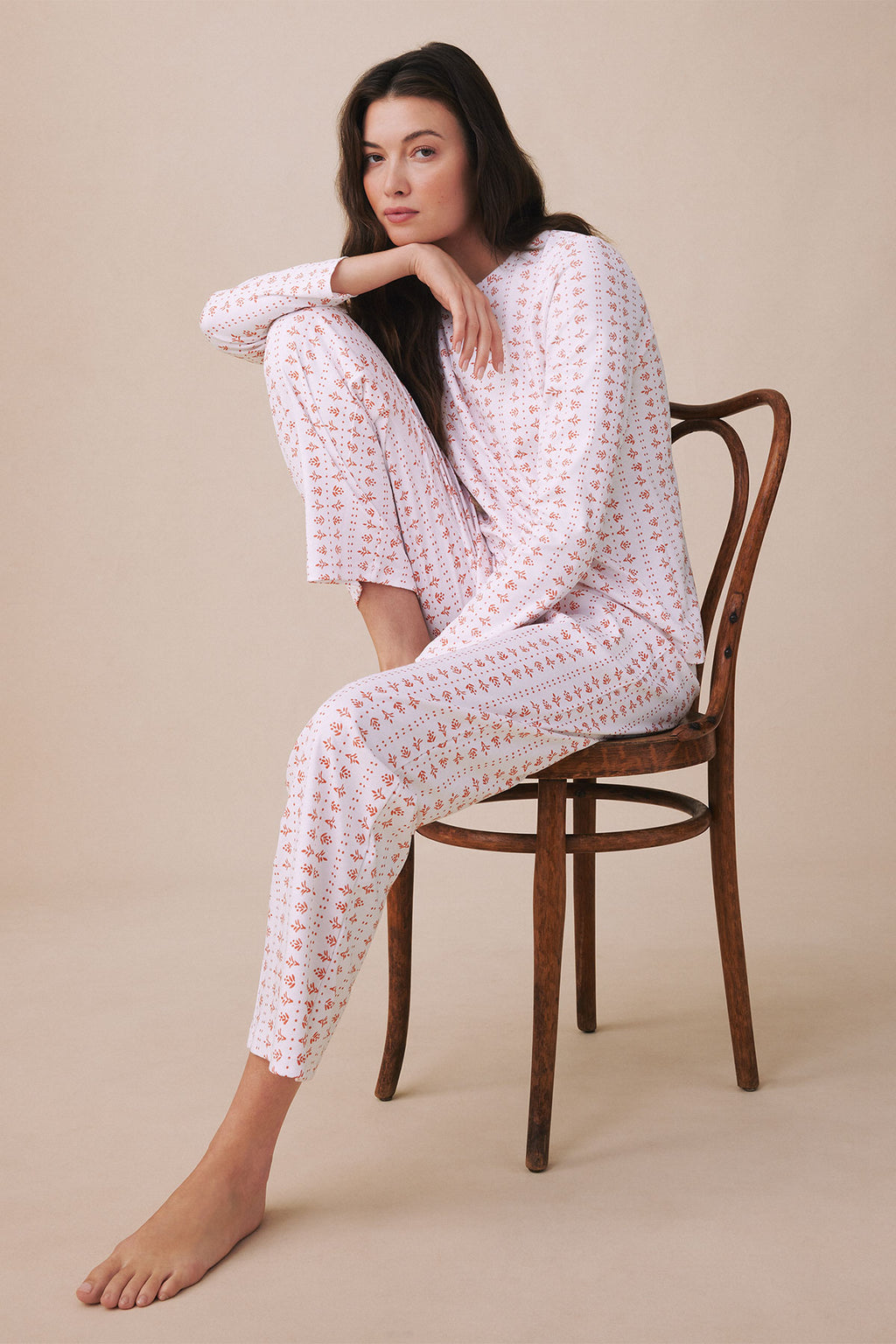 Woman wearing white long-sleeve pajama set with small red floral print, seated on wooden chair.