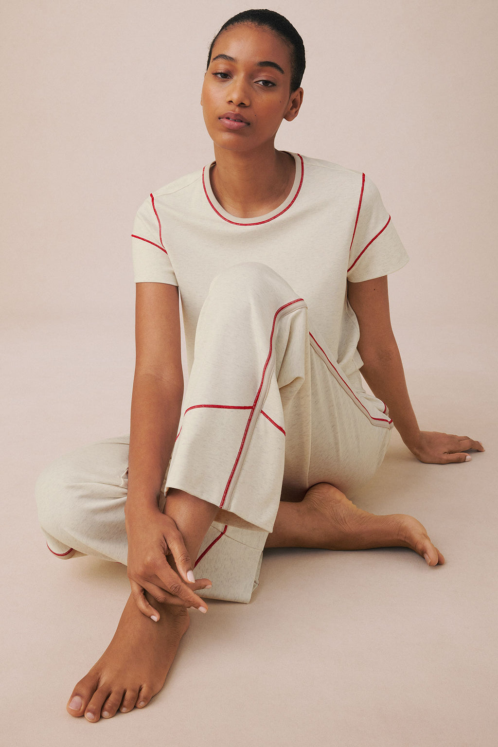 Cream-colored short-sleeve top and cropped pants set with red contrast piping, worn by model seated on neutral background.