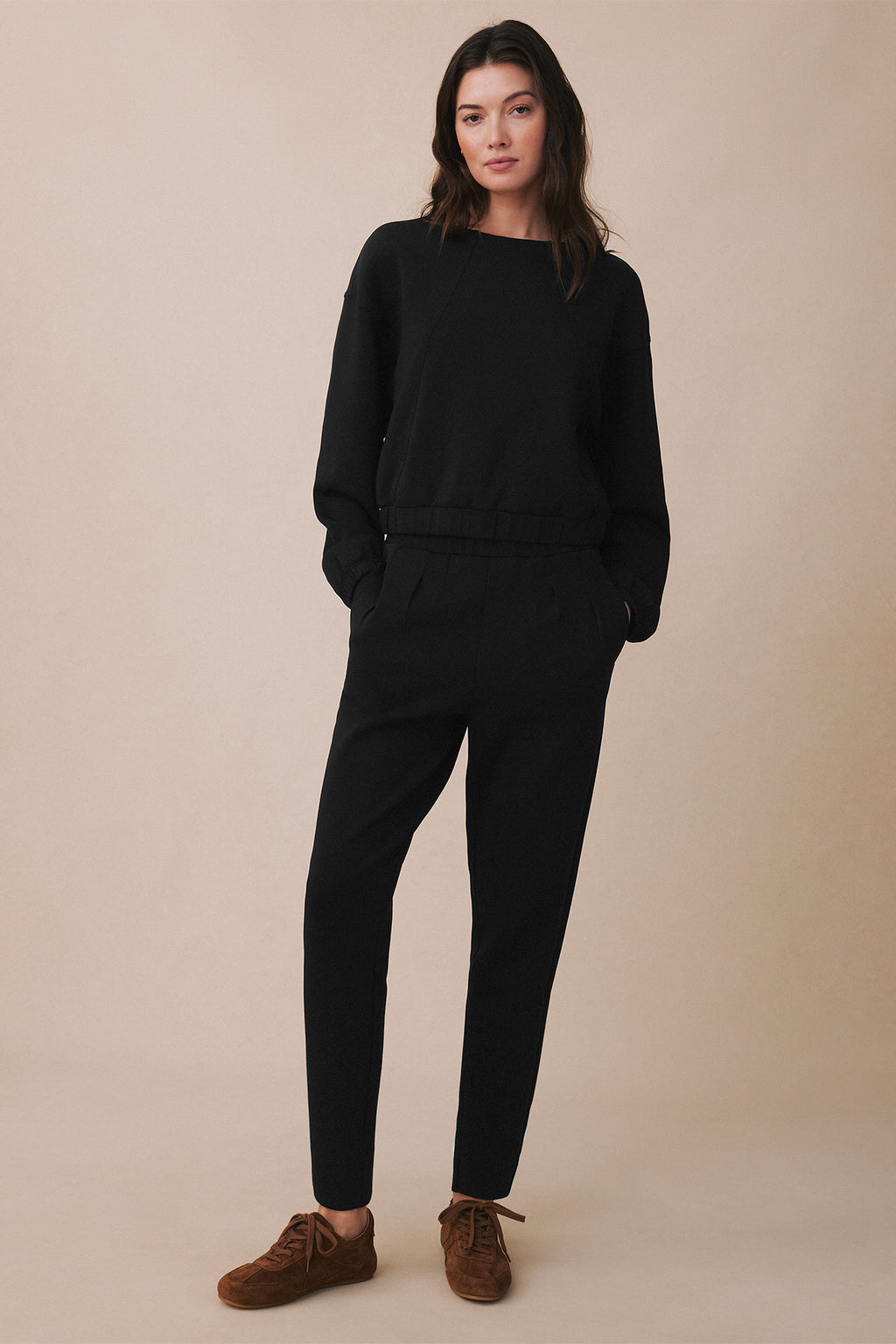 Woman wearing black long-sleeved jumpsuit with tapered legs and brown suede shoes against beige background.