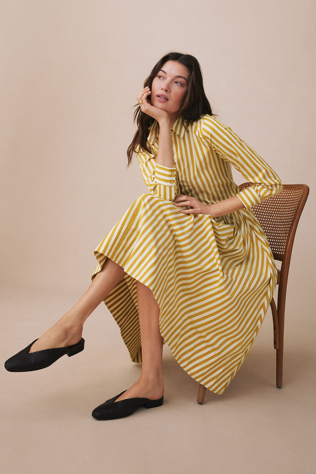 Woman wearing yellow and white striped button-up shirt and matching skirt with black slip-on shoes, seated on a woven chair.