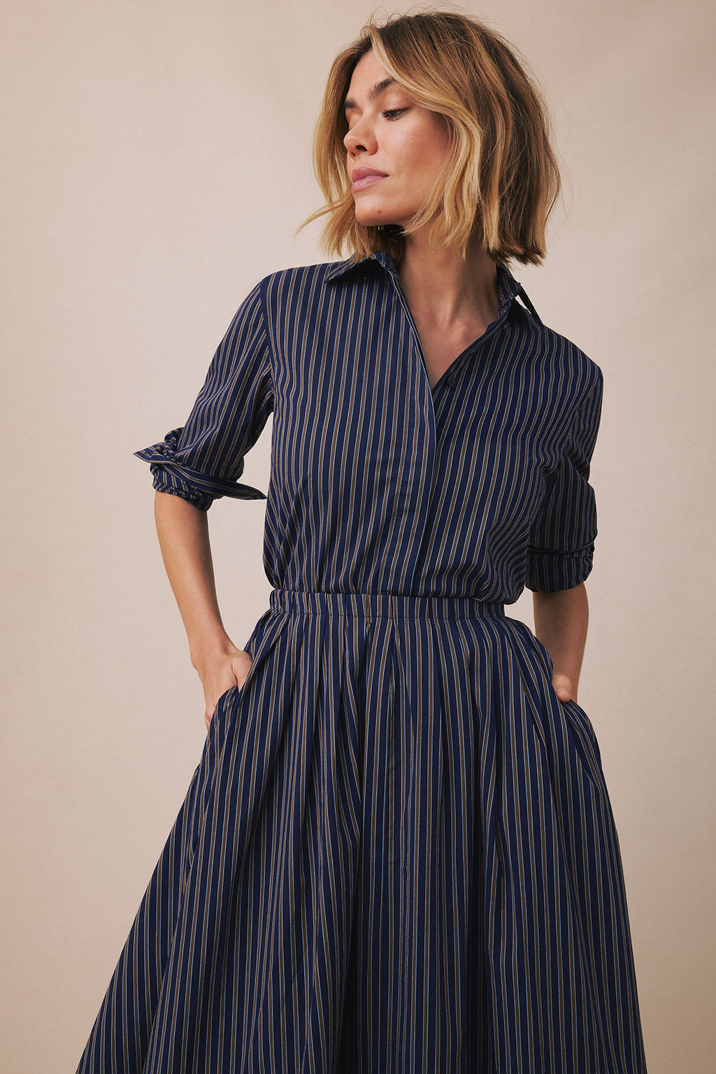 Woman wearing navy and white striped button-up shirt with matching pleated skirt and side pockets.