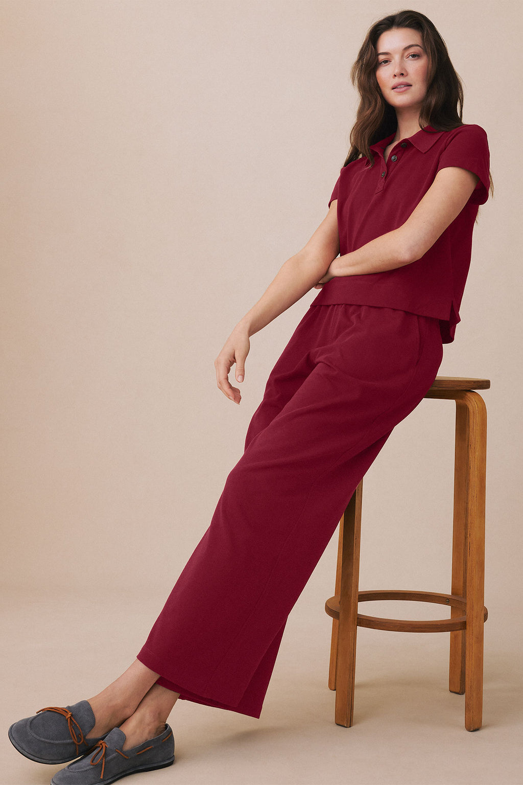 Woman wearing burgundy polo shirt and matching wide-leg pants, seated on wooden stool.