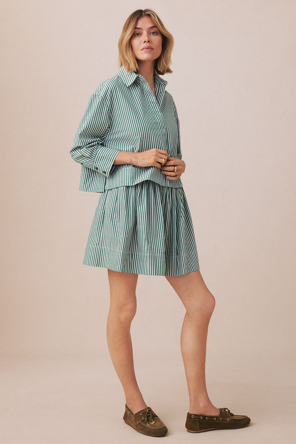 Woman wearing hunter green and white striped button-up shirt and matching pleated skirt with brown loafers.
