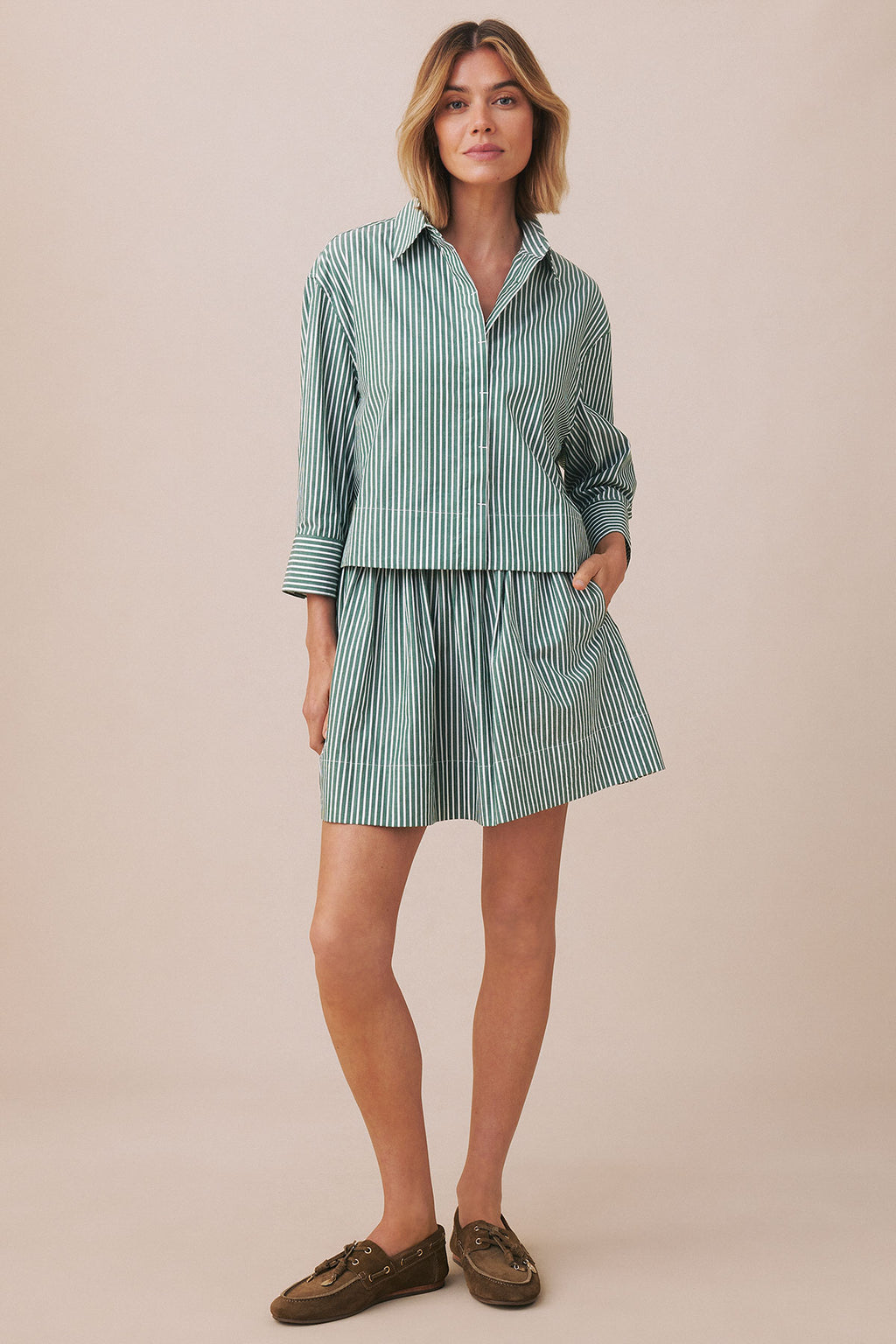 Woman wearing a hunter green and white vertical striped button-up shirt and matching skirt set with brown loafers.