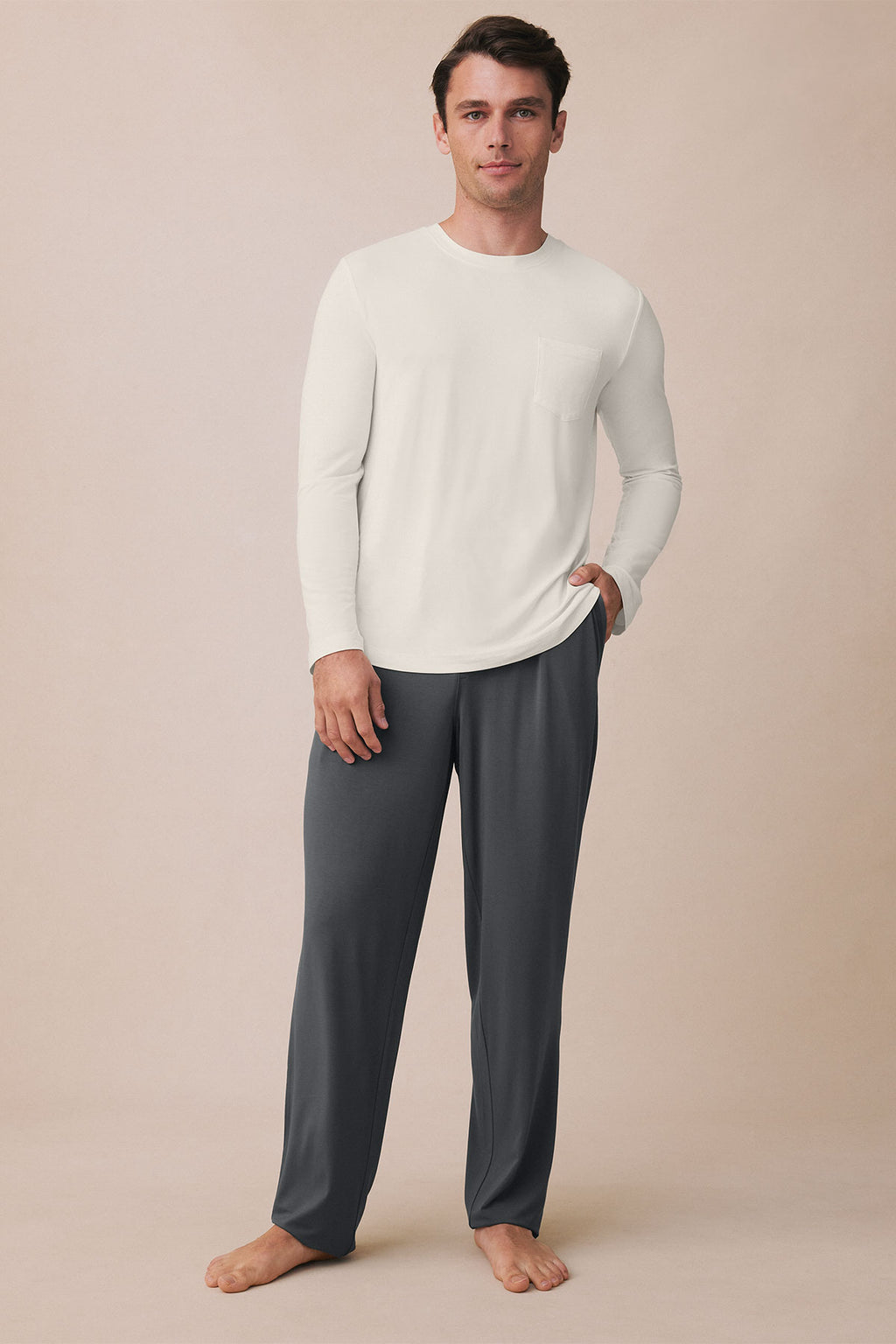 Man wearing white long-sleeve shirt and charcoal pants standing against neutral background.