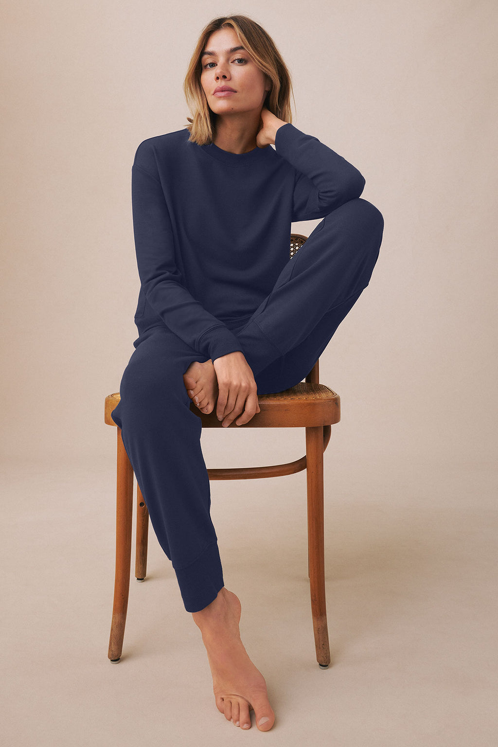 Woman wearing navy long-sleeve crewneck sweatshirt and matching jogger pants, seated on wooden chair.