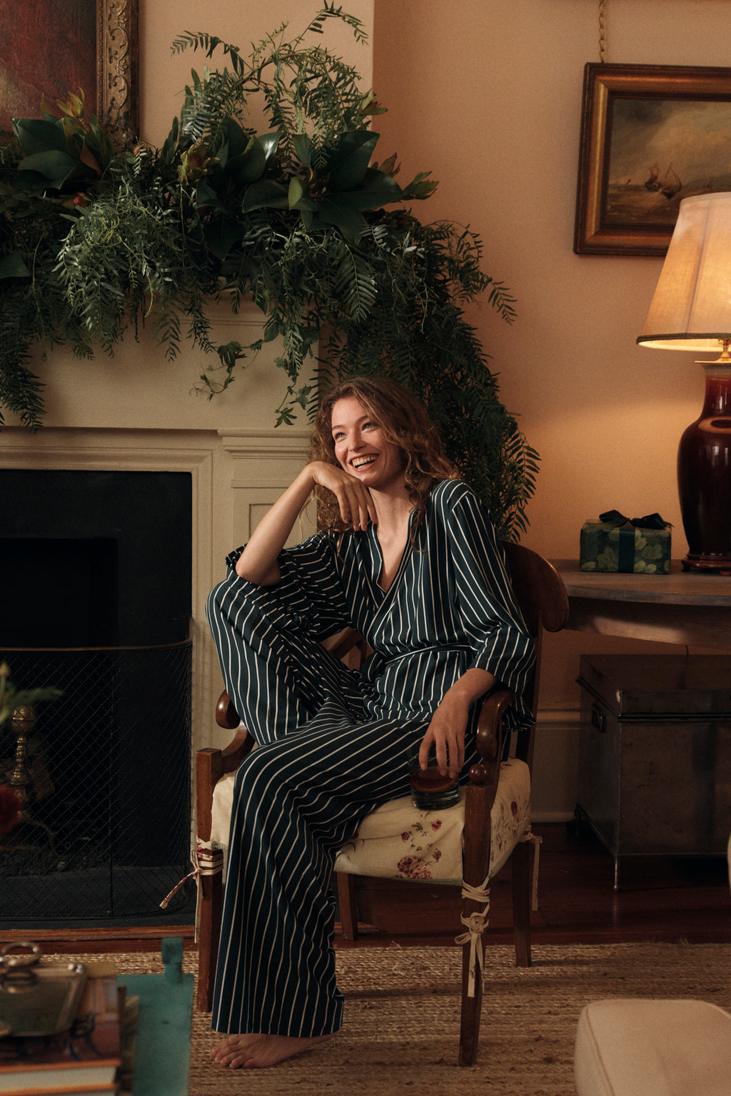 A woman wears the DreamModal Kimono Pajama Set in Conifer Stripe, smiling as she relaxes on a floral armchair. Cozy decor, greenery above a fireplace, and soft table lamp lighting create a warm, inviting atmosphere around her.