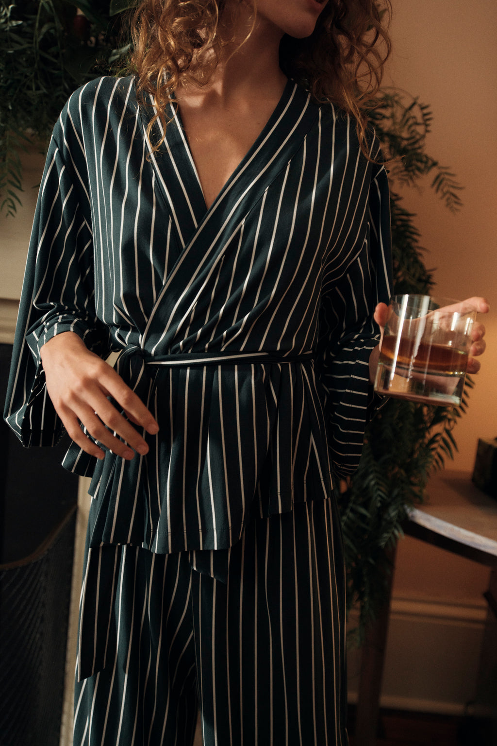 Wearing the DreamModal Kimono Pajama Set in Conifer Stripe, a person holds a glass of water. Cozy indoor lighting and lush green plants enhance the inviting atmosphere in the background.