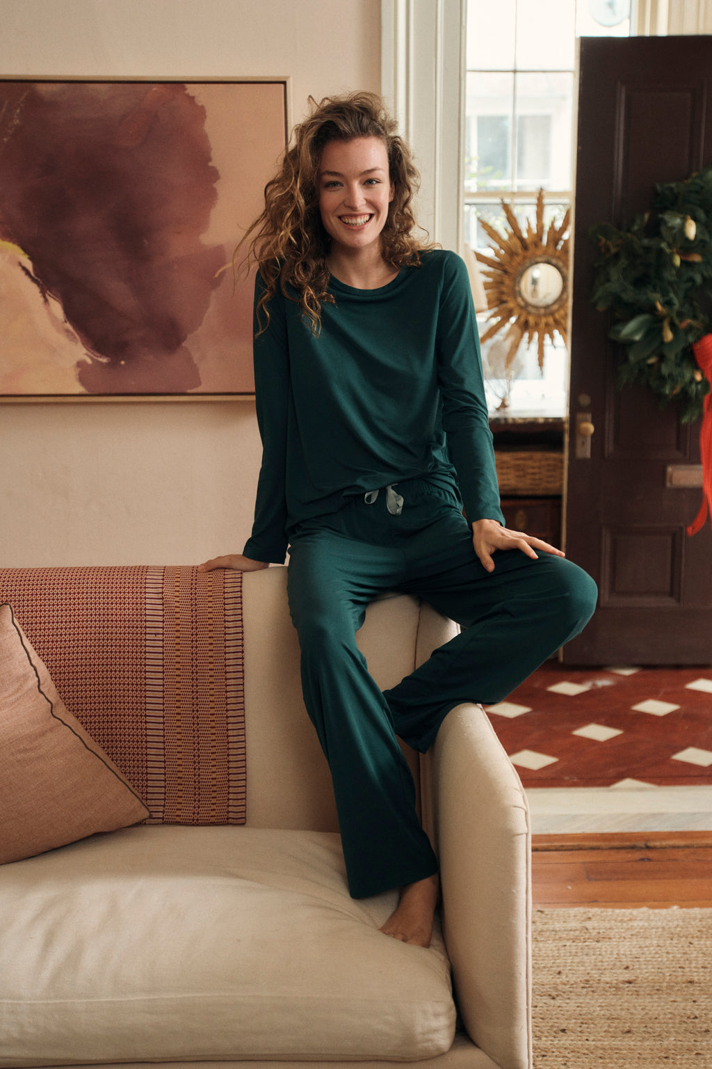 A woman in the DreamModal Ribbon Long-Long Set in Conifer sits smiling on the arm of a beige couch in a cozy living room, decorated with a holiday wreath and abstract artwork.