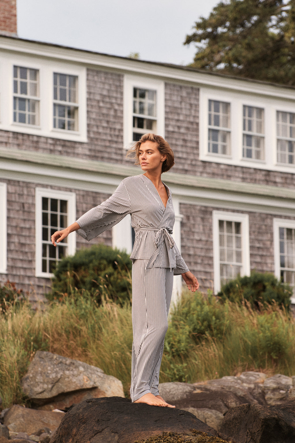Wearing the DreamModal Kimono Pajama Set in Black Pencil Stripe, a person stands on large rocks outdoors, extending one arm and gazing into the distance, with a gray shingled house and greenery in the background.